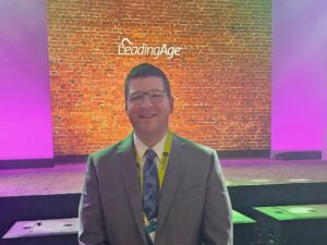 A smiling Michael Y., Health Services Administrator, celebrates his recent graduation from the esteemed Larry Minnix Leadership Academy. The backdrop glows with fuchsia and purple lighting that pops against the warm orange hues of a brick wall.